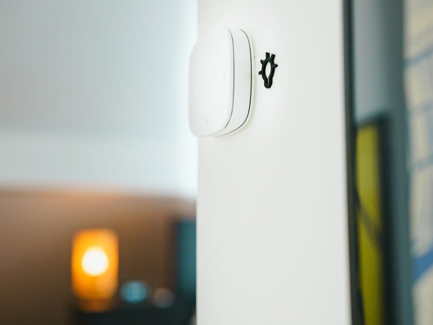 Commande d'éclairage connectée fixée au mur d'un salon, une lampe allumée en arrière-plan